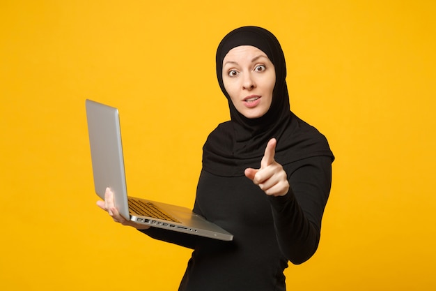 Jeune femme musulmane arabe en vêtements noirs hijab tenir et travailler un ordinateur portable isolé sur un portrait de mur jaune. Concept de mode de vie religieux des gens.