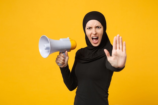 Jeune femme musulmane arabe en vêtements noirs hijab tient dans la main un mégaphone d'adresse publique isolé sur un portrait de mur jaune. Concept de mode de vie religieux des gens.