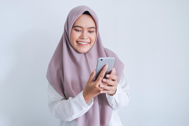 Jeune femme musulmane asiatique portant un foulard est souriante et heureuse de ce qu'elle voit sur le smartphone Femme indonésienne sur fond gris