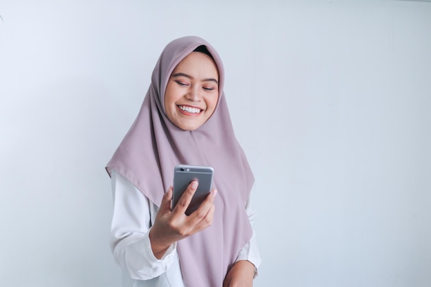 Jeune femme musulmane asiatique portant un foulard est souriante et heureuse de ce qu'elle voit sur le smartphone Femme indonésienne sur fond gris