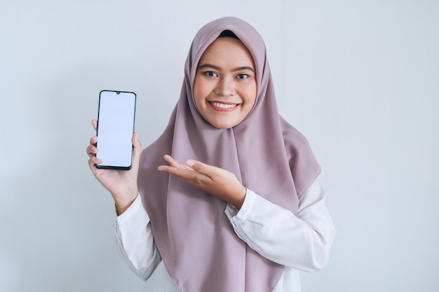 Jeune femme musulmane asiatique portant un foulard pointe le doigt sur l'écran blanc du téléphone avec le sourire et le sentiment heureux Femme indonésienne sur fond gris