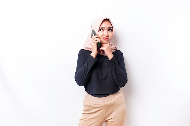 Une jeune femme musulmane asiatique réfléchie porte le hijab et semble confuse entre les choix tout en parlant sur son téléphone isolé par un fond blanc