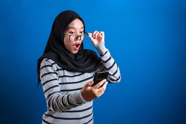 Jeune femme musulmane expression choquée, regardant son téléphone sur fond bleu