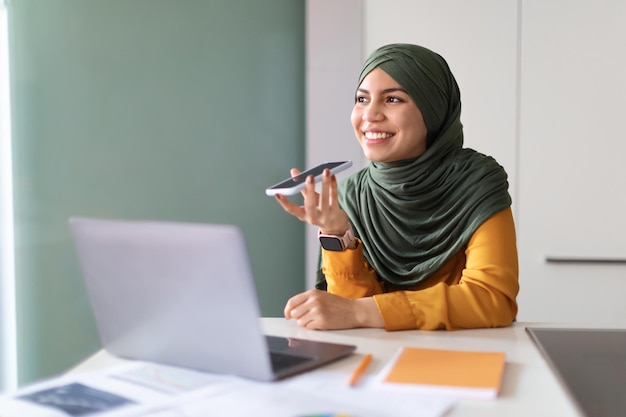 Jeune femme musulmane en hijab enregistrant un message vocal sur un smartphone au bureau