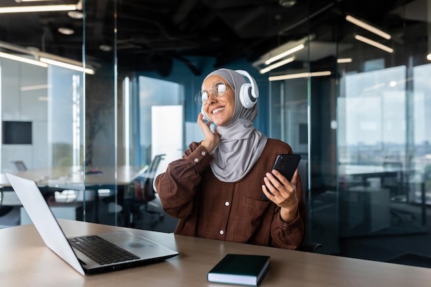 Une jeune femme musulmane en hijab est assise au bureau au bureau et écoute de la musique dans