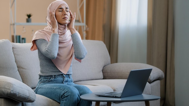 Jeune femme musulmane en hijab rose assise sur un canapé travaillant à l'aide d'un ordinateur portable ayant des maux de tête migraine triste