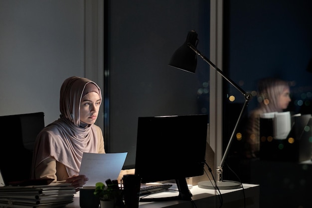 Jeune femme musulmane en hijab tenant du papier et regardant un écran d'ordinateur