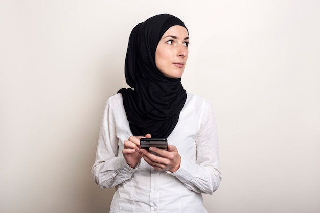 Jeune femme musulmane en hijab tient un téléphone dans ses mains et regarde sur le côté