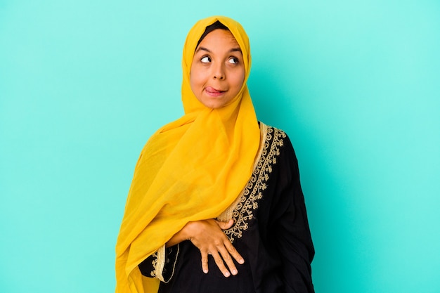 Jeune femme musulmane isolée sur le mur bleu touche le ventre, sourit doucement, concept de manger et de satisfaction.