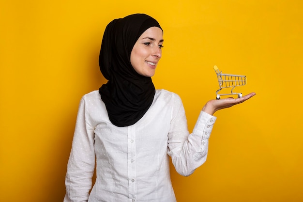 Jeune femme musulmane joyeuse en hijab tenant un caddie sur un jaune