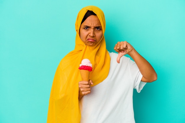 Jeune femme musulmane mangeant une glace isolée sur un mur bleu montrant un geste d'aversion, les pouces vers le bas. Concept de désaccord.