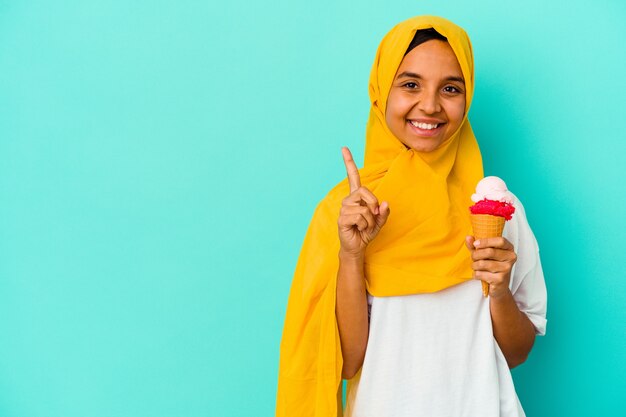 Jeune femme musulmane mangeant une glace isolée sur un mur bleu montrant le numéro un avec le doigt.