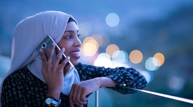 Jeune femme musulmane portant un voile d'écharpe sur la rue de la ville urbaine la nuit envoyant des SMS sur un smartphone avec la lumière de la ville bokeh en arrière-plan