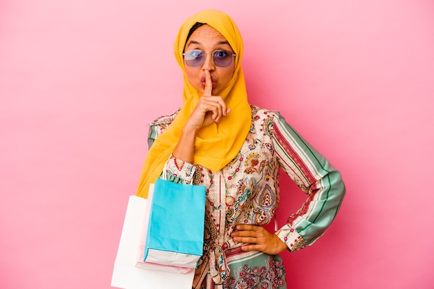 Jeune femme musulmane shopping des vêtements isolés sur un mur rose en gardant un secret ou en demandant le silence