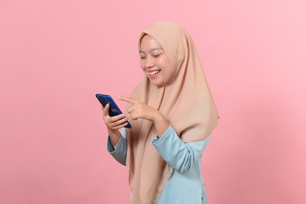 Une jeune femme musulmane souriante et souriante tient un téléphone intelligent dans les mains, en regardant des messages texte sur l'écran d'affichage à l'aide d'applications de médias sociaux en ligne isolées sur fond rose