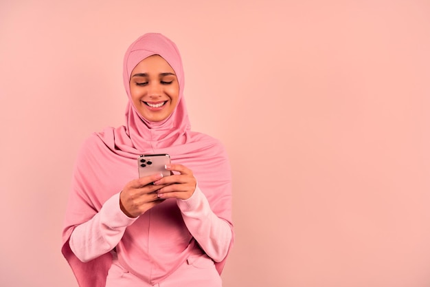 Une jeune femme musulmane souriante en vêtements rose tendre regarde le téléphone et sourit
