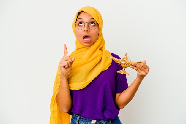 Jeune femme musulmane tenant une lampe isolée sur fond blanc pointant vers le haut avec la bouche ouverte.