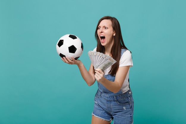 Une jeune femme nerveuse qui crie fan de football soutient l'équipe préférée avec un ballon de football, fan d'argent en billets de banque en dollars en espèces isolé sur fond de mur bleu turquoise. Concept de loisirs sportifs en famille.