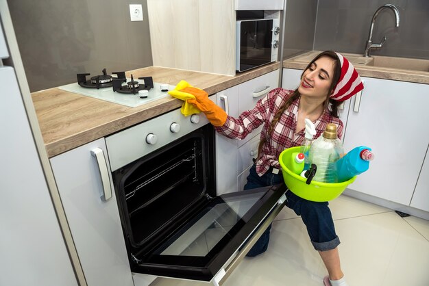 Jeune femme nettoyant le four avec un chiffon jaune et un spray dans la cuisine.