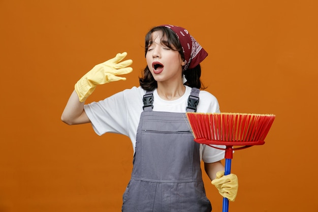 Jeune femme nettoyante concentrée portant des gants en caoutchouc uniformes et un bandana tenant une vadrouille à raclette en gardant la main dans l'air avec les yeux fermés se préparant à éternuer isolé sur fond orange