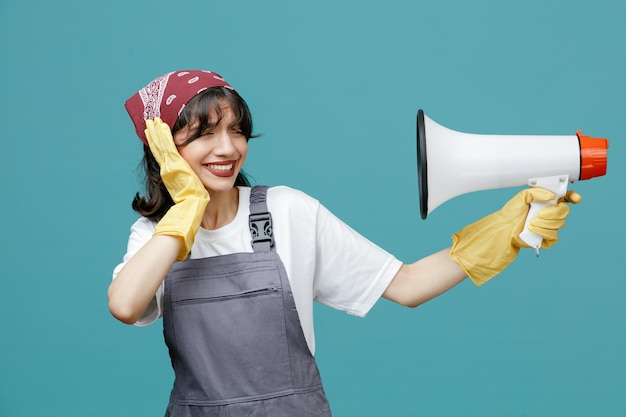 Jeune femme nettoyeuse irritée portant un bandana uniforme et des gants en caoutchouc pointant le haut-parleur vers elle-même en le regardant fermer l'oreille avec la main isolée sur fond bleu