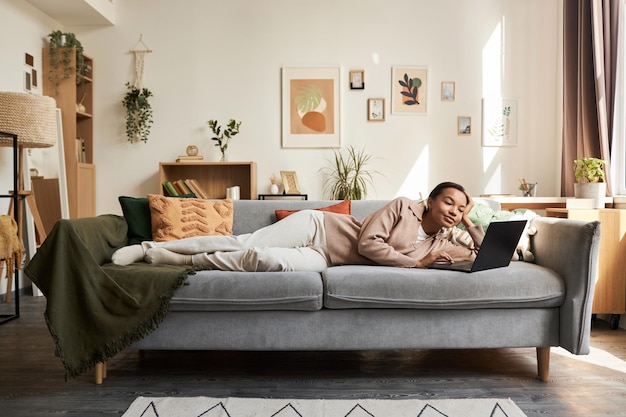 Une jeune femme noire allongée sur le canapé dans un intérieur confortable et utilisant un ordinateur portable