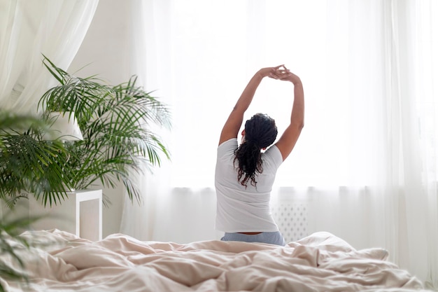 Jeune femme noire assise dans son lit et étirant les bras après s'être réveillée