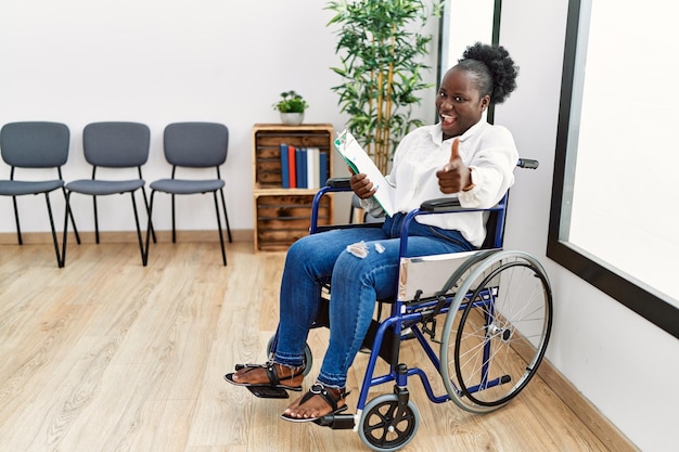 Jeune femme noire assise sur un fauteuil roulant dans la salle d'attente, pointant du doigt la caméra avec un visage heureux et drôle. bonne énergie et vibrations.