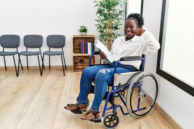 Jeune femme noire assise sur un fauteuil roulant dans la salle d'attente souriante gaie montrant et pointant avec les doigts les dents et la bouche concept de santé dentaire