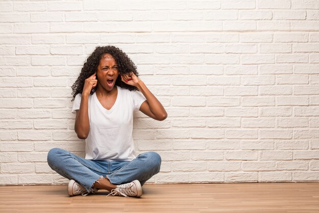 Jeune femme noire assise sur un plancher en bois, couvrant les oreilles avec les mains, en colère et fatiguée d&#39;entendre du son