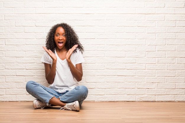 Jeune femme noire assise sur un plancher en bois surpris et choqué, regardant avec de grands yeux