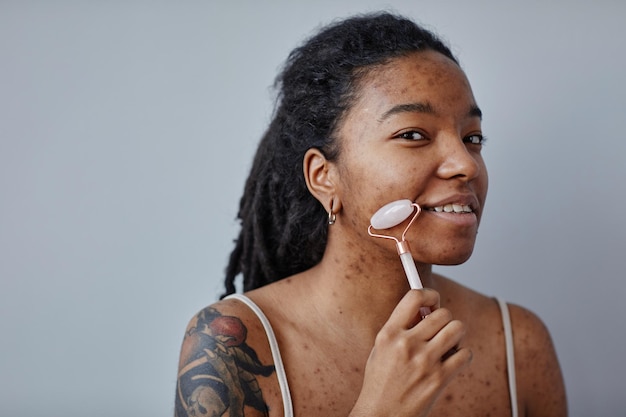 Jeune femme noire avec des cicatrices d'acné et des tatouages à l'aide d'un masseur de visage