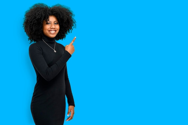 Jeune femme noire avec une coiffure afro sourit et pointe vers un espace sur le côté droit, elle porte une robe noire, un espace pour le texte ou le produit, concept de promotion