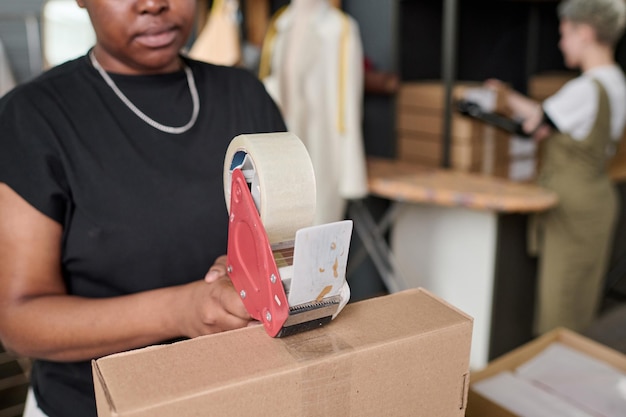 Photo jeune femme noire avec du ruban adhésif scellant une boîte en carton avec des marchandises emballées