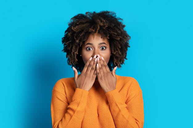 Une jeune femme noire émotionnelle surprise se couvre la bouche en regardant la caméra