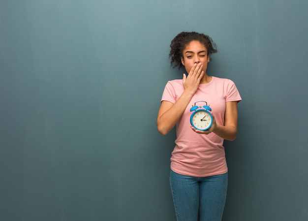 Jeune femme noire fatiguée et très endormie. Elle tient un réveil.