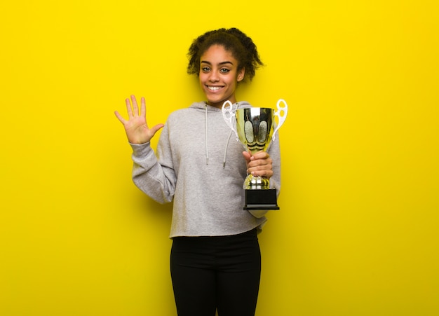 Jeune femme noire de fitness montrant le numéro cinq. Tenant un trophée.