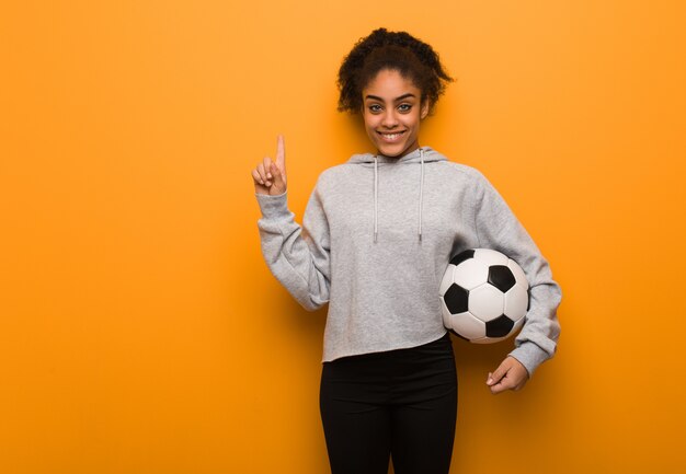 Jeune femme noire fitness montrant le numéro un. Tenir un ballon de foot.