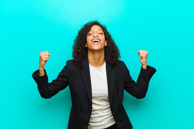 Jeune femme noire heureuse, surprise et fière, criant et célébrant le succès avec un grand sourire contre le mur bleu