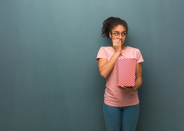 Jeune femme noire se ronger les ongles, nerveuse et très inquiète. Elle tient un seau de maïs soufflé.