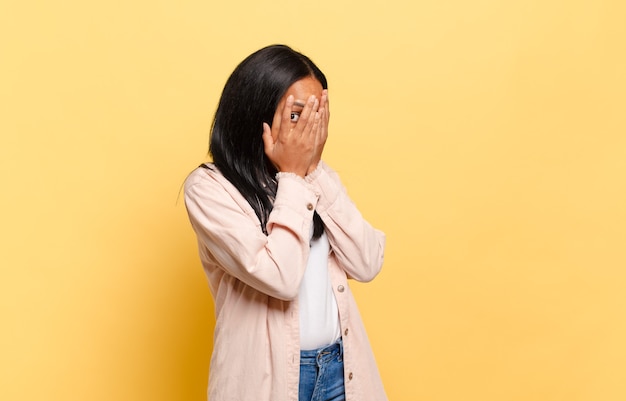 Jeune femme noire se sentant effrayée ou gênée, jetant un coup d'œil ou espionnant avec les yeux à moitié couverts de mains