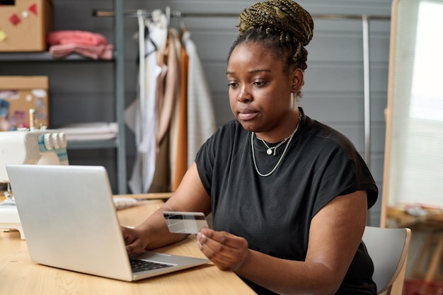 Jeune femme noire sérieuse avec carte de crédit assise devant un ordinateur portable