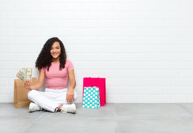 Jeune femme noire souriant joyeusement avec une main sur la hanche et une attitude confiante, positive, fière et amicale assise sur le sol