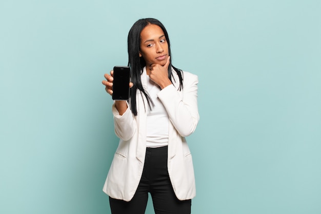 Jeune femme noire souriante avec une expression heureuse et confiante avec la main sur le menton, se demandant et regardant sur le côté. concept de téléphone intelligent