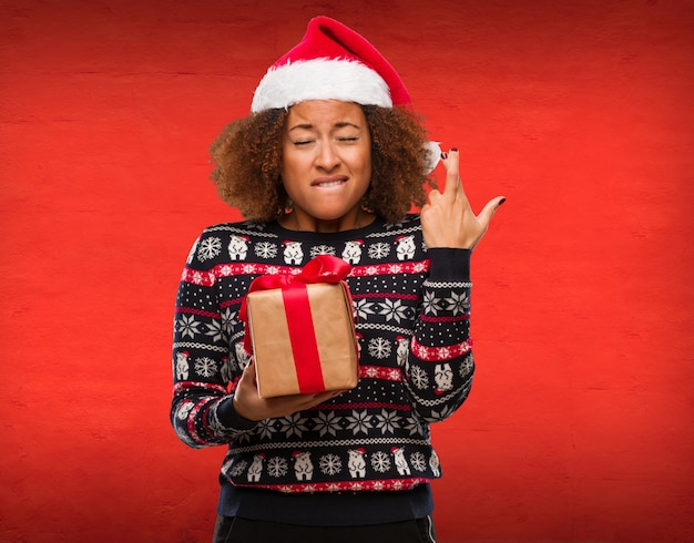 Jeune femme noire tenant un cadeau au jour de Noël, croise les doigts pour avoir de la chance