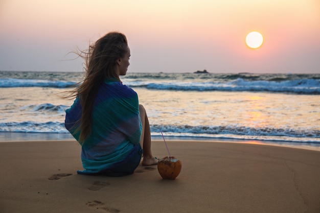 La jeune femme avec une noix de coco regarde le coucher du soleil