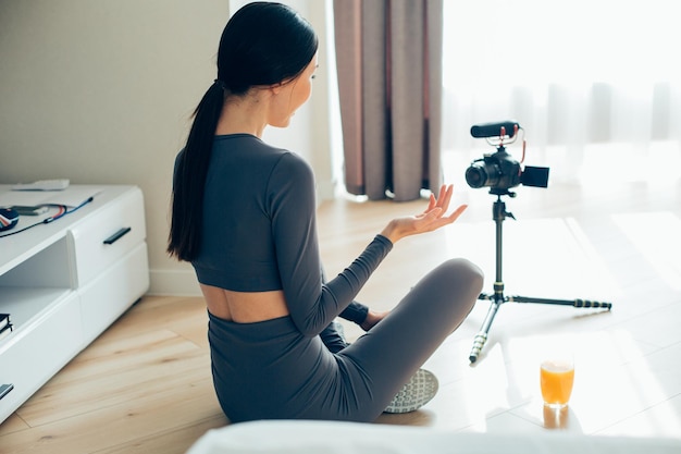 Jeune femme non reconnue en tenue de sport assise sur le sol à la maison devant une caméra sur trépied