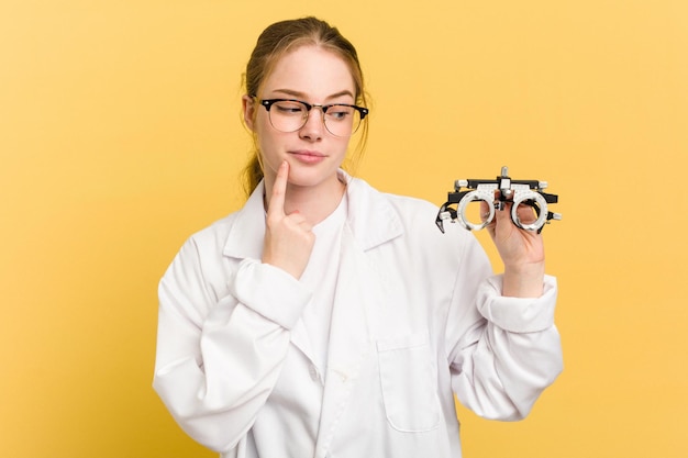 Jeune femme oculiste caucasienne tenant des lunettes d'optométrie isolées sur fond jaune