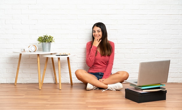 Jeune femme avec un ordinateur portable assis sur le sol à l'intérieur heureux et souriant coning bouche avec la main