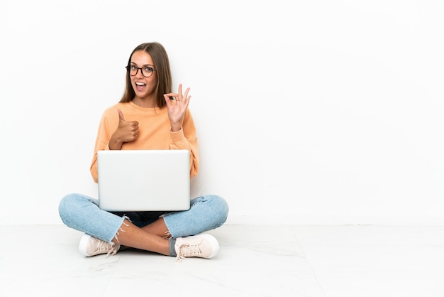 Jeune femme avec un ordinateur portable assis sur le sol montrant signe ok et geste de pouce vers le haut